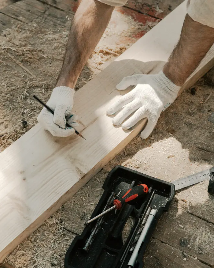 Gloved hands marking a cut line on a pine board with a carpenter's pencil