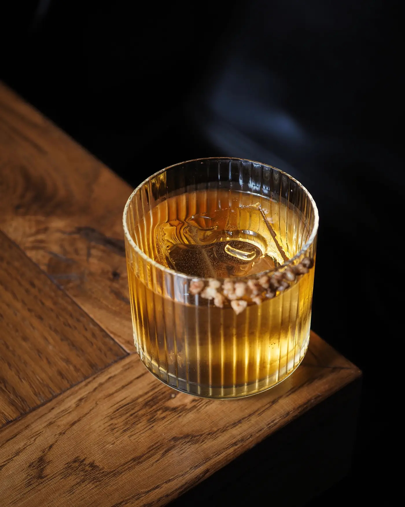 A whiskey cocktail in a ribbed glass on the corner of a wood bar