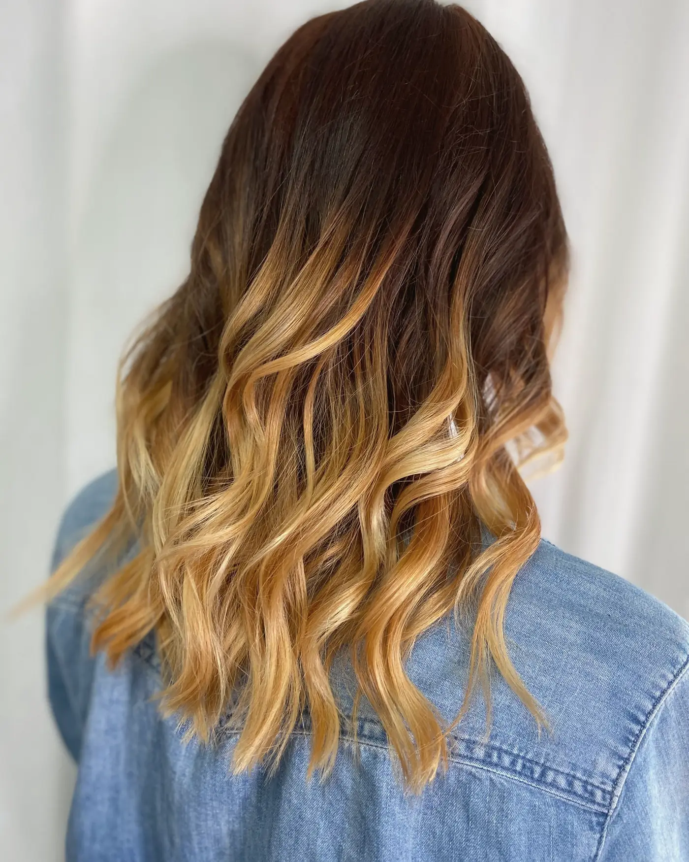 Brunette hair with a soft golden balayage, seen from behind against a white wall
