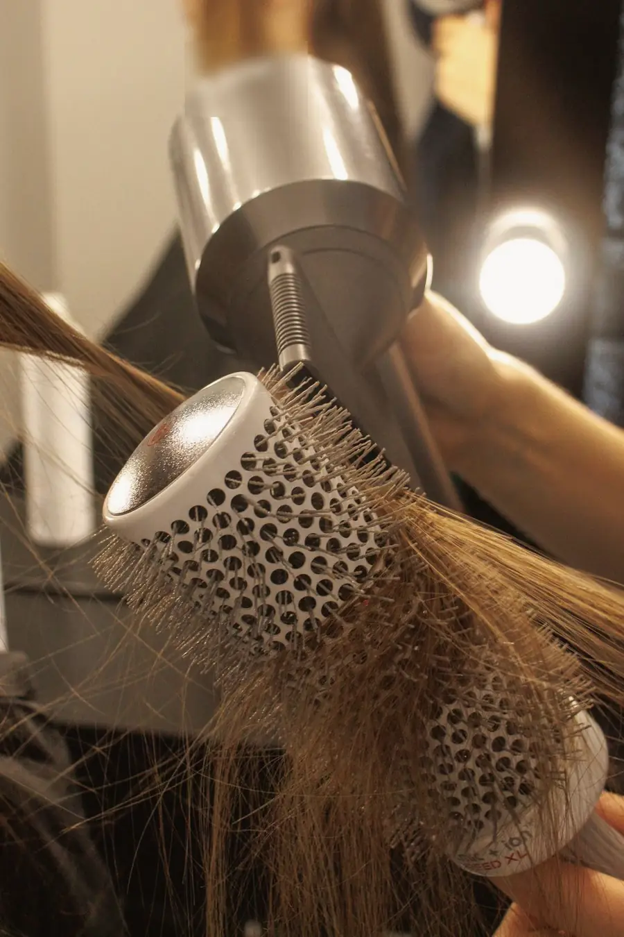 A stylist blow-drying long brown hair with a round brush under mirror lights